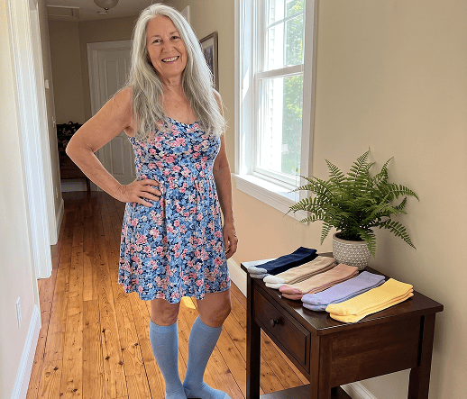 Woman standing in a floral dress wearing pale blue compression socks, with folded colorful sock pairs displayed nearby